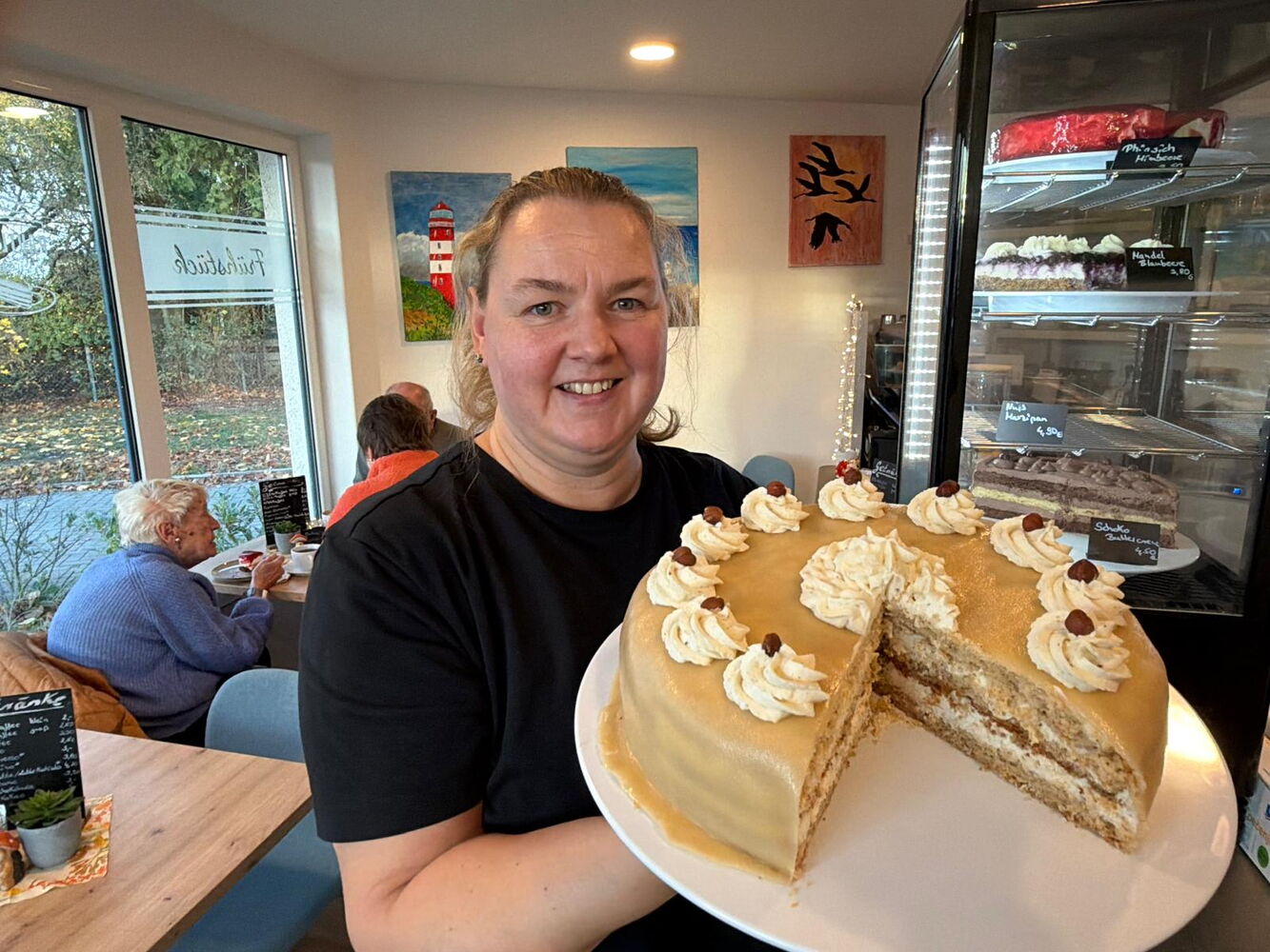 Frau hält Torte im Café, im Hintergrund Gäste und Kunst.