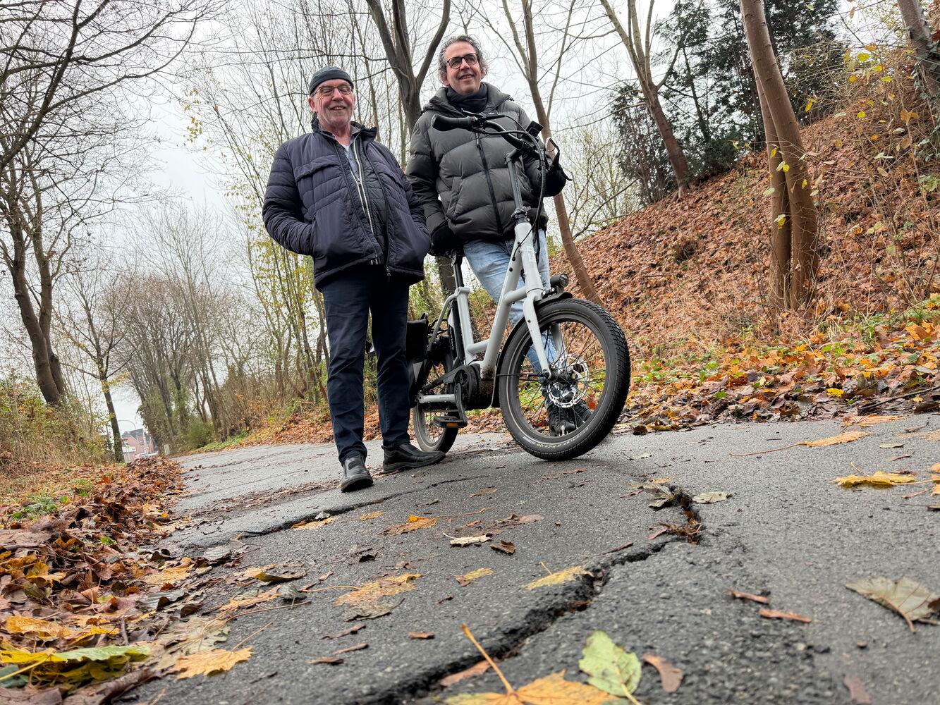 Zwei Männer posieren mit Fahrrad auf herbstlichem Weg.