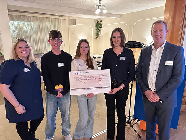 Fünf Personen halten einen großen Scheck in einem Raum.