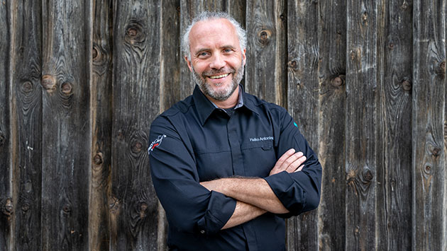 Mann lächelt vor einer Holzwand mit verschränkten Armen.