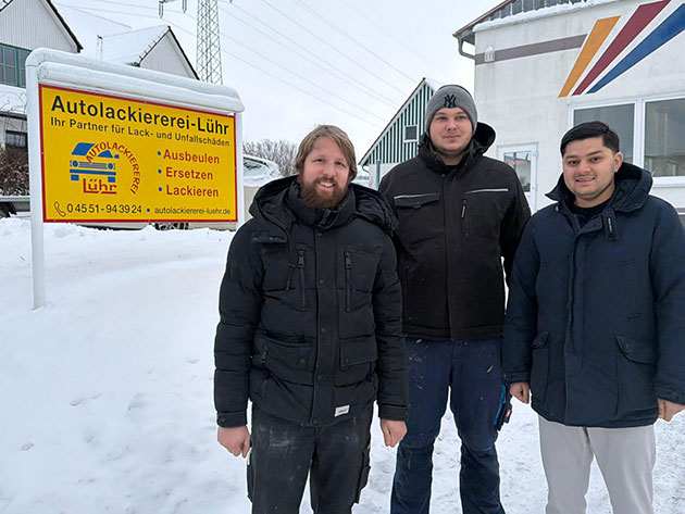 Drei Männer vor Autolackiererei-Schild im Schnee.
