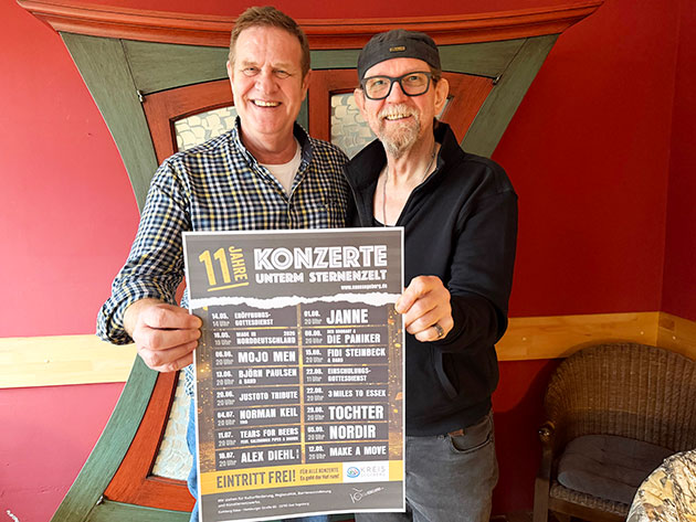 Zwei Männer halten ein Konzertplakat in einem Raum.
