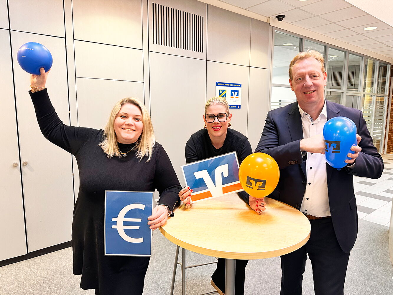 Drei Personen mit Ballons und Logos einer Bank.