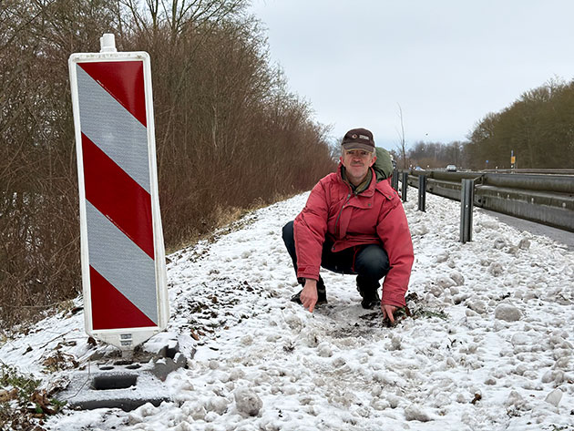 Mann kniet im Schnee neben einer Leitplanke und einem Warnschild