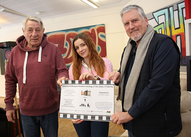 Drei Personen halten ein großes Poster mit Veranstaltungsdetails.