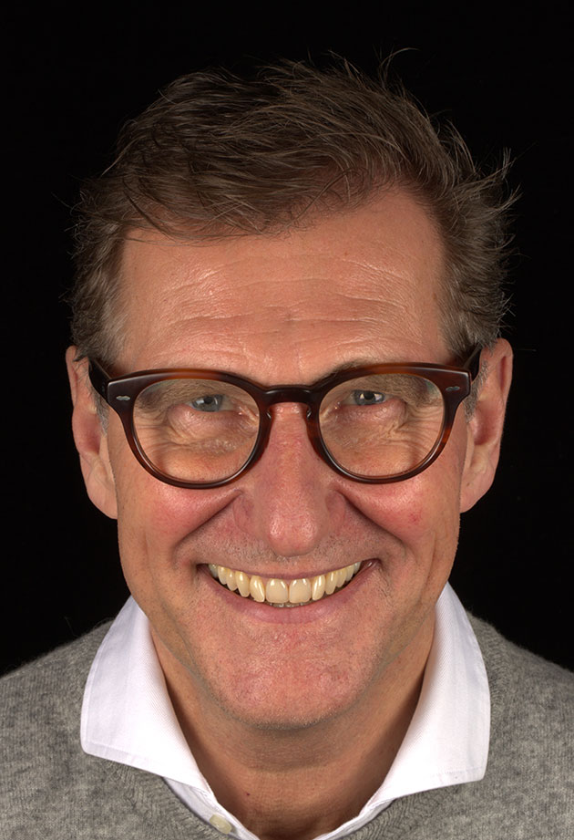 Lächelnder Mann mit Brille in grauem Pulli vor schwarzem Hintergrund.