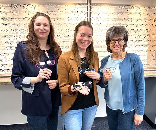Drei Frauen zeigen verschiedene Brillen vor Brillenregal.