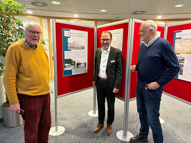 Drei Männer vor rot hinterlegten Infotafeln in einem Raum.