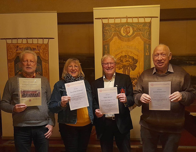 Vier Personen halten Urkunden vor gemusterten Bannern.