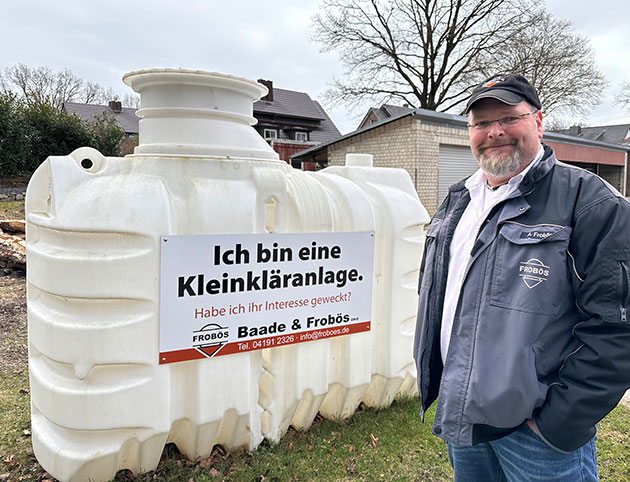 Mann steht neben einer weißen Kleinkläranlage mit Schild.
