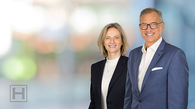 Zwei Personen in Businesskleidung lächeln vor unscharfem Hintergrund.