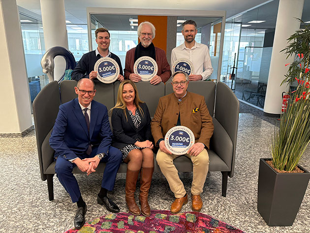 Sechs Personen, drei halten 3000€-Schilder in einem Büro.