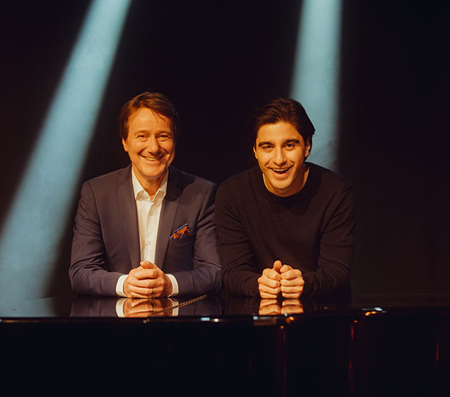 Zwei Männer lächeln im Scheinwerferlicht hinter einem Klavier.
