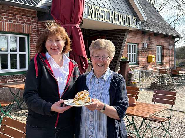 Zwei Frauen halten Kuchen vor einem Backsteinhaus.