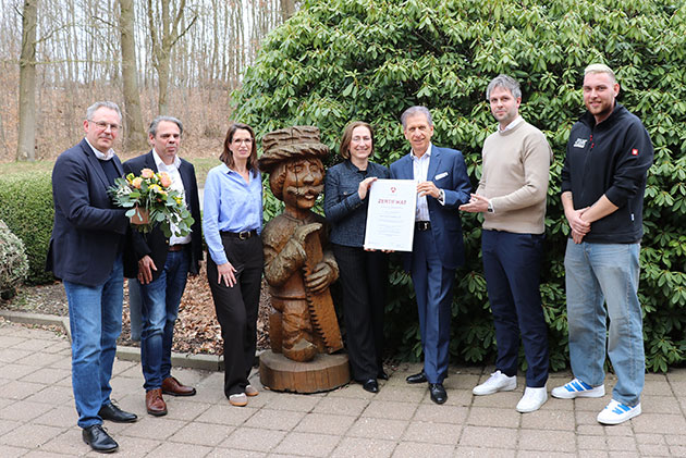 Gruppe posiert mit Zertifikat neben Holzfigur im Freien.