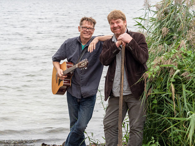 Zwei Männer am Seeufer, einer mit Gitarre.