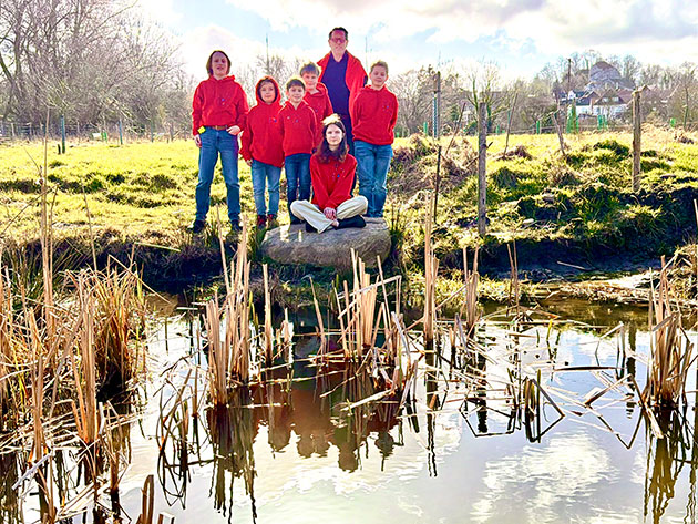 Gruppe steht am Teichrand, sonniger Tag, Schilf im Vordergrund.