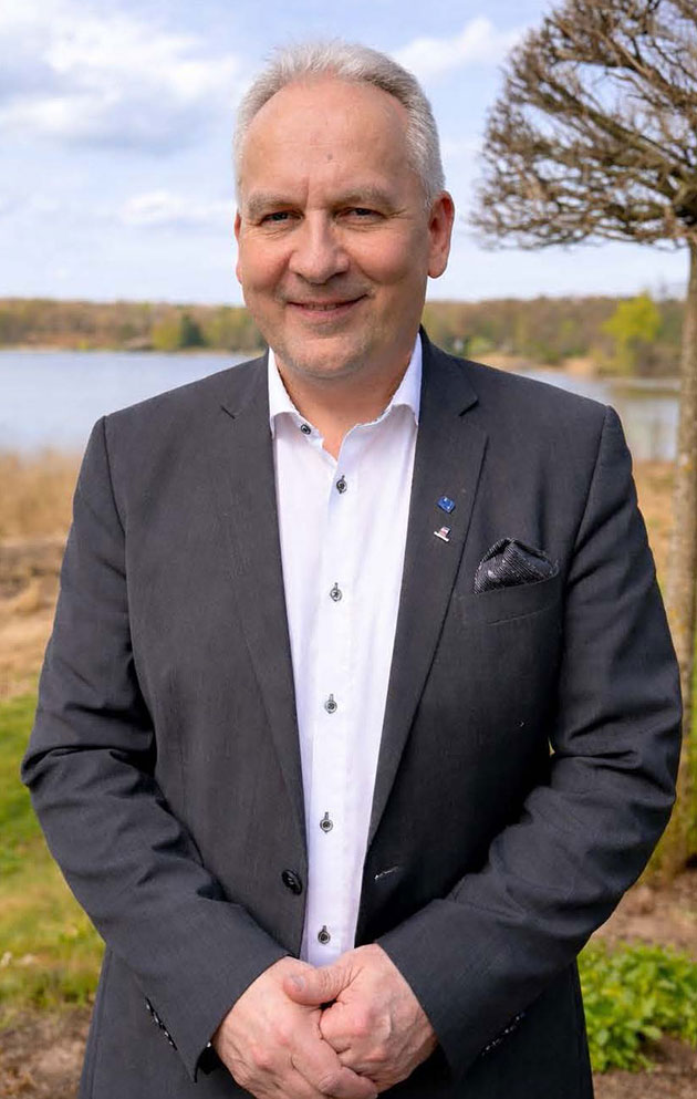 Mann im Anzug lächelt im Freien vor einem See.