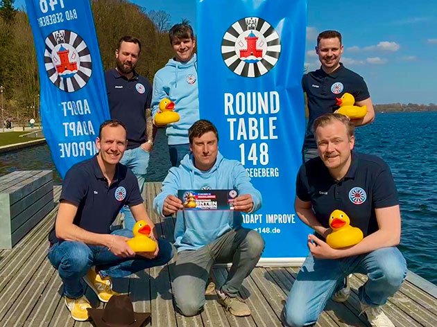 Gruppe von Männern mit Enten an einem See vor Bannern.