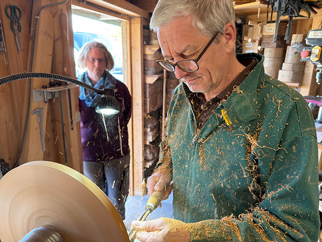 Mann drechselt Holz in der Werkstatt, eine Person schaut zu.