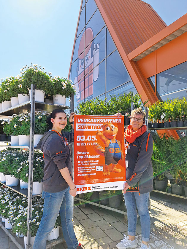 Zwei Personen halten ein Poster vor einem Baumarkt.
