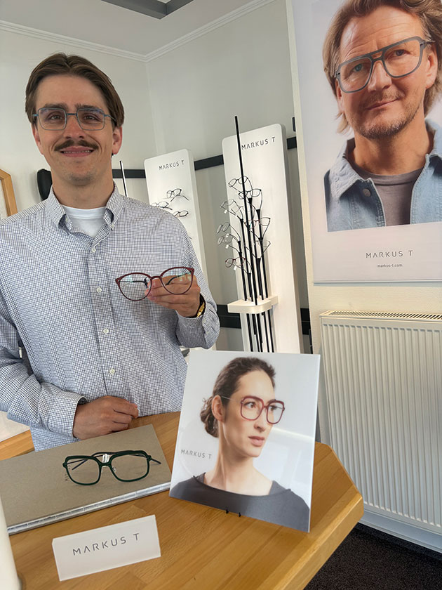 Mann hält Brille in Optiker-Geschäft mit Plakaten.