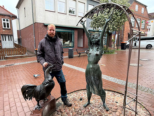 Mann steht neben Bronzeskulpturen auf einer regennassen Straße.