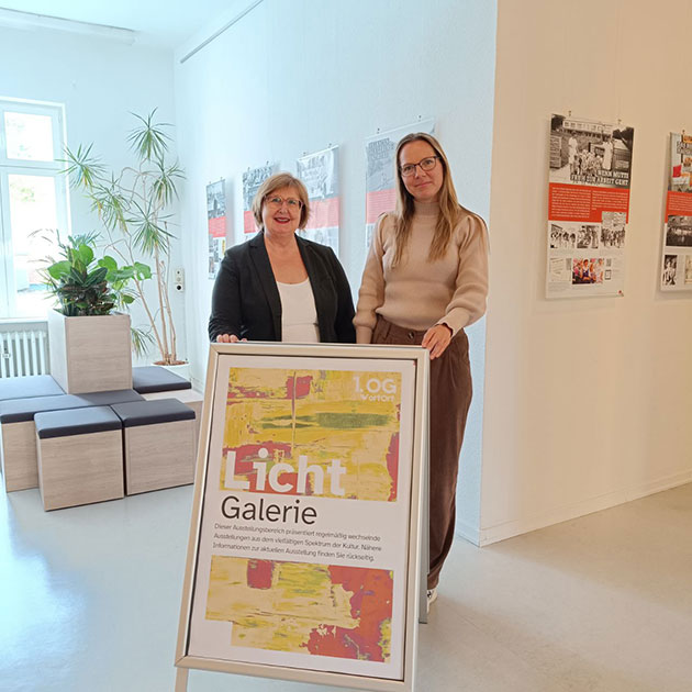 Zwei Frauen posieren neben einem Galerieschild in einem Ausstellungsraum.