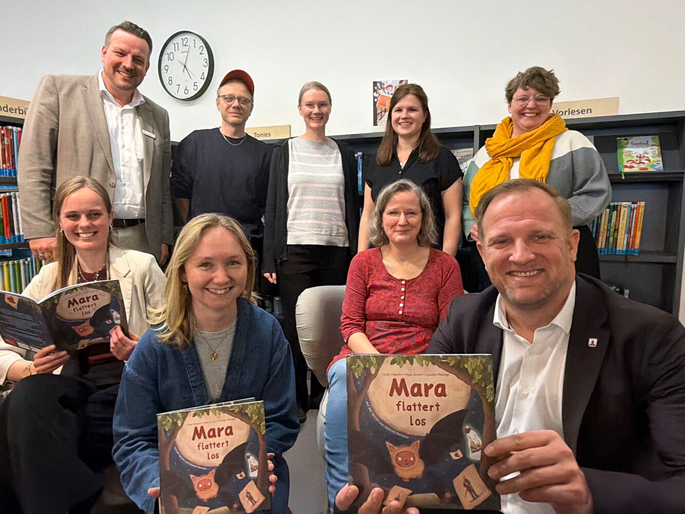 Gruppe posiert mit dem Buch 'Mara flattert los' in einer Bibliothek.