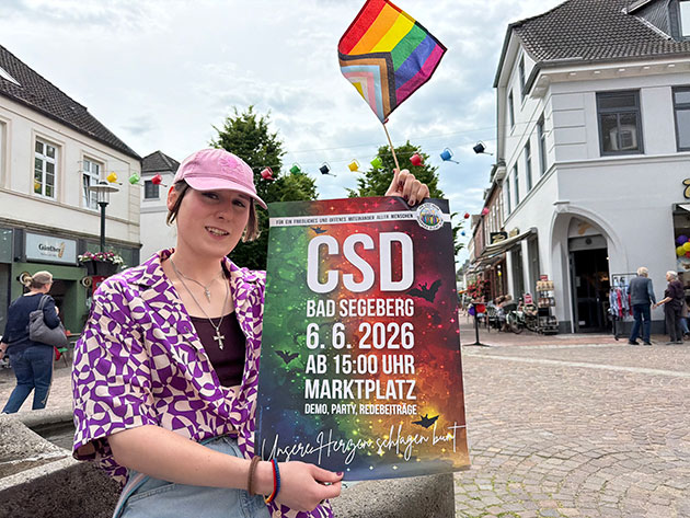 Person hält CSD-Plakat auf einem Stadtplatz.