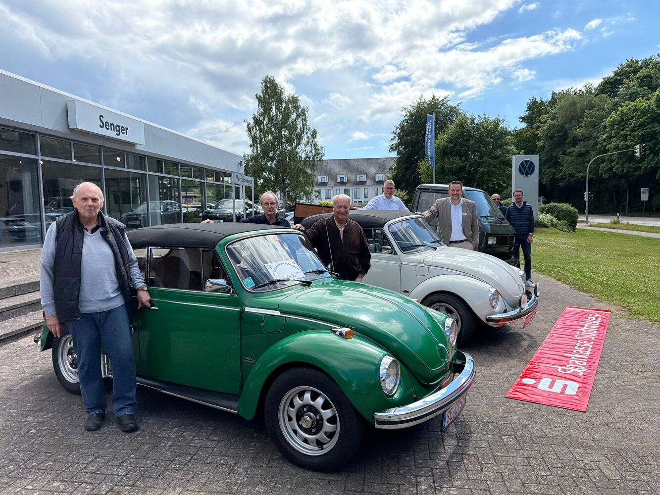 Gruppe posiert mit klassischen VW-Käfern vor Autohaus.