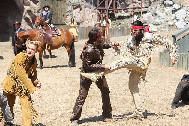 Szene aus Western-Show mit Cowboys und Indianern auf Sandboden.
