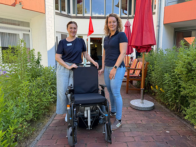 Zwei Frauen stehen neben einem Rollstuhl im Freien.
