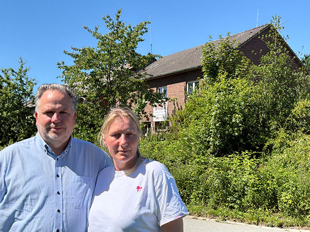 Zwei Personen stehen vor einem überwucherten Backsteinhaus.