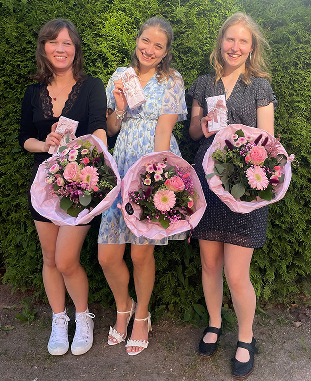 Drei Frauen mit Blumensträußen und Karten lächeln vor einer Hecke.