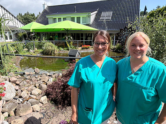 Zwei Krankenschwestern vor einem Gartenteich mit Haus im Hintergrund.