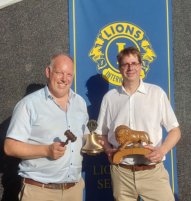 Zwei Männer mit Objekten vor einem Lions Club Banner.