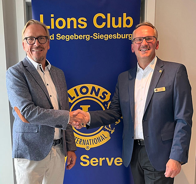 Zwei Männer schütteln sich vor einem Lions Club Banner die Hand.