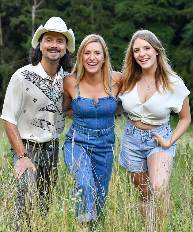Drei Personen lachen auf einer Wiese in sommerlicher Kleidung.