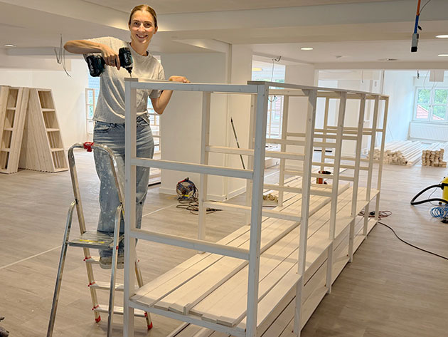 Lächelnde Person baut ein weißes Regal mit Bohrmaschine zusammen.