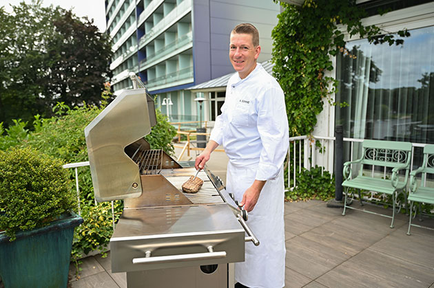 Koch grillt Fleisch auf einer Terrasse vor einem Gebäude.