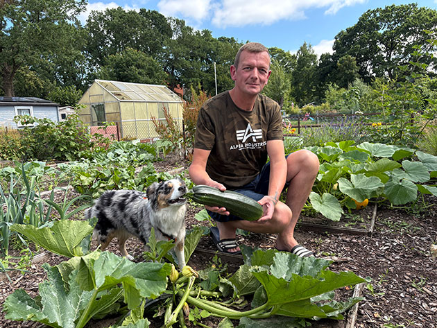 Mann mit Zucchini und Hund im Garten vor Gewächshaus