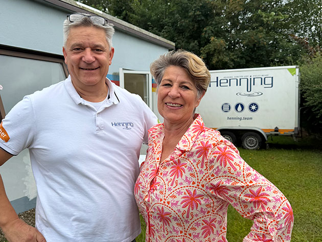 Zwei Personen lächeln vor einem Gebäude mit Henning-Logo.