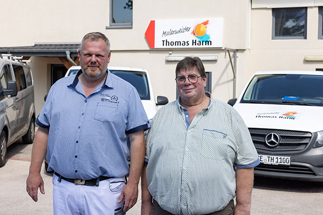 Zwei Männer vor einem Malergeschäft mit Autos im Hintergrund.