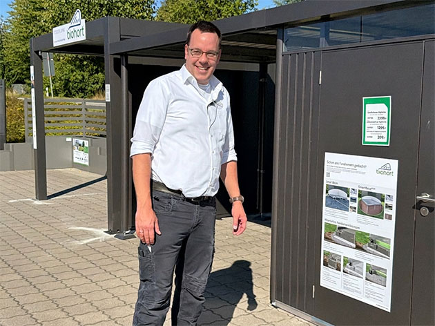 Mann in weißem Hemd vor einem Gartenhaus auf einem Parkplatz