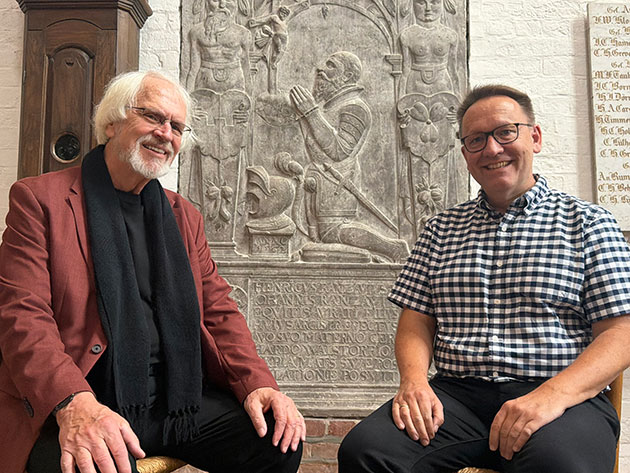 Zwei Männer sitzen vor einem historischen Relief in der Kirche.