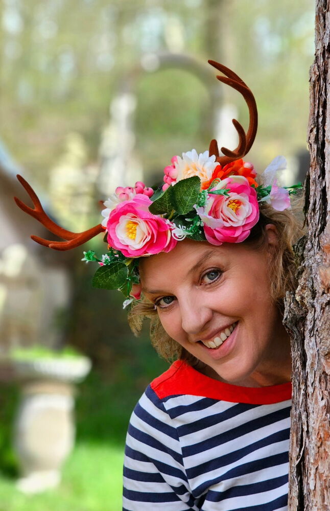 Frau mit Blumen- und Geweihkrone lächelt hinter einem Baum hervor.