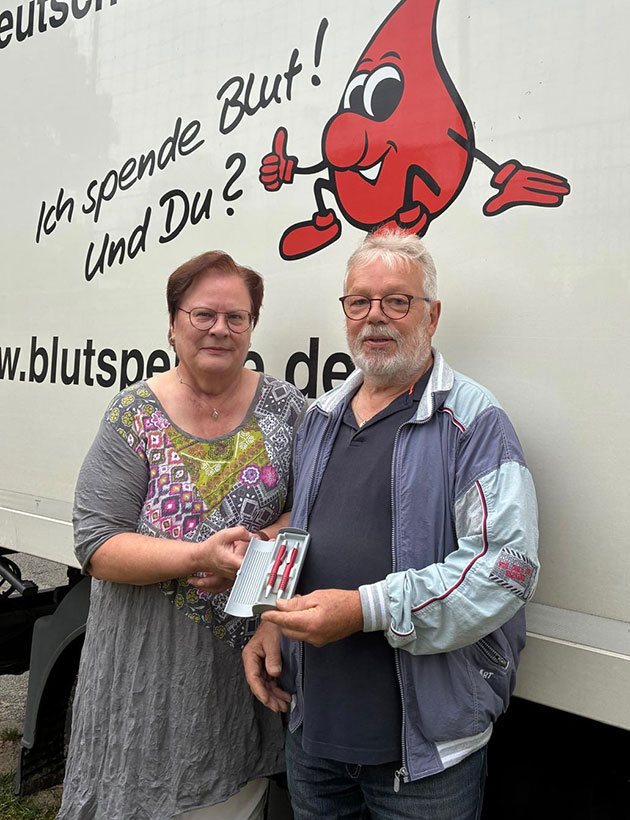 Zwei Personen vor einem Blutspende-Lkw mit Geschenk.