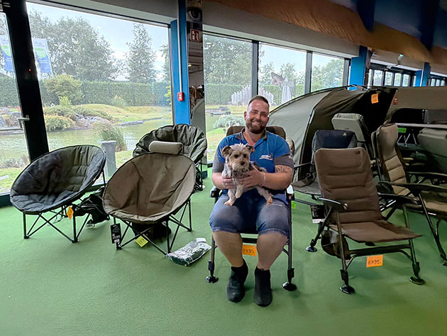 Mann mit Hund auf Stuhl in Campingladen, Zelte im Hintergrund.