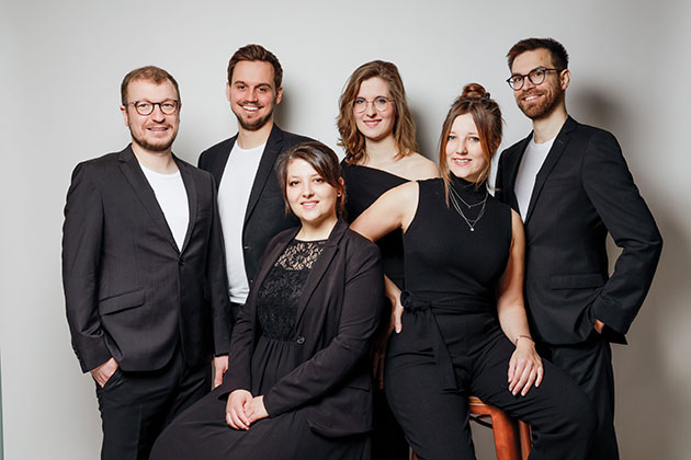 Sechs Personen in eleganter Kleidung posieren zusammen.